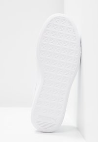 Semelle d’une basket blanche Puma avec un motif en forme d’étoile, inclinée contre un mur blanc d’angle.