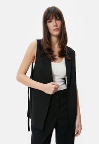 Schwarzer ärmelloser Blazer mit tailliertem Design, ausgestattet mit einem einzelnen Knopfverschluss und seitlichen Bindebändern, getragen über einem weißen gerippten Tanktop.