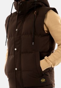 Brown padded vest with a hood, featuring snap buttons, side pockets, and drawstring accents. Worn over a beige knitted sweater.