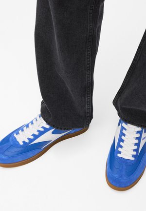 Modré semišové a syntetické tenisky s bílými akcenty, které mají strukturovanou podrážku a bílé tkaničky, spárované s tmavými džínovými kalhotami.