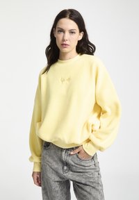 Geel fleece sweatshirt met ronde hals, geribbelde mouwen en geborduurd logo op de borst. Draagt het met grijze high-waisted spijkerbroek.