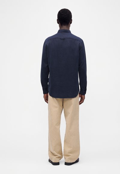 Man stands facing away wearing a navy long-sleeve shirt, beige wide-leg pants, and black shoes against a plain white background.