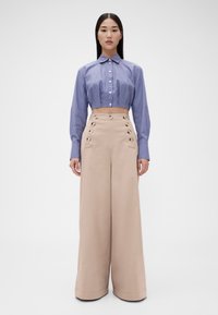 Lyhyt nappipaita sinivalkoisilla raidoilla, yhdistetty korkeavyötäröisiin, leveälahkeisiin beigeihin housuihin, joissa on nappikoristeet sivuilla.