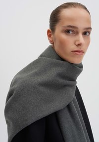 Una sciarpa di lana grigia si adagia elegantemente attorno al collo, caratterizzata da una consistenza morbida e una finitura liscia, completata da un cappotto scuro sottostante.
