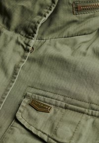 Groene visgraatstof, close-up van een ritsvak en een metalen logo-badge, met getextureerde stikdetails en een knoopsluiting.