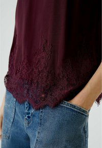 Burgunderbluse mit Spitzenbesatz am Saum. Der Stoff ist leicht und glatt. Kombiniert mit blauer Denim im klassischen Schnitt mit Tasche.