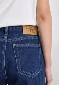 Tamsiai mėlynos džinsinės kelnės klasikinio kirpimo, su šviesiai rudos odos juosmens etikete, kurioje įspausta "Calvin Klein Jeans", ir užpakaliniu kišeniu.