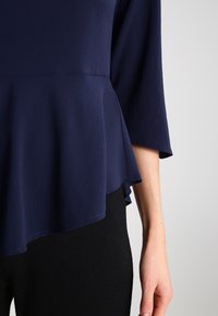 Top peplum blu navy con una texture liscia e maniche a tre quarti. L'orlo è leggermente curvo, sovrapposto a pantaloni neri.