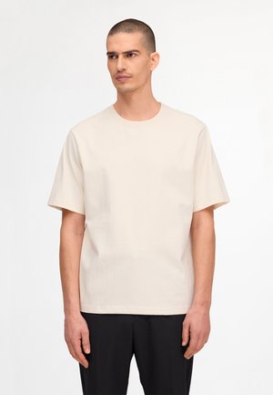 Mężczyzna o krótkich włosach, ubrany w jednolity beżowy t-shirt z krótkim rękawem i czarne spodnie, stoi na białym tle.