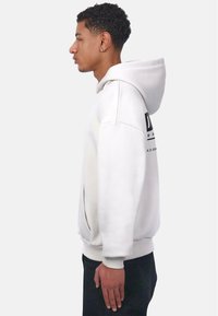 Sweatshirt à capuche blanche avec une poche kangourou, arborant un texte graphique noir au dos, porté avec un pantalon sombre, vu de côté.