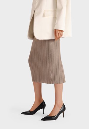 Donna con gonna midi beige a coste, blazer crema e décolleté nere con punta, in piedi su sfondo bianco.