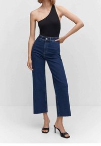 Bodysuit negro de un solo hombro combinado con jeans de denim azul oscuro de tiro alto y pierna ancha. La modelo lleva sandalias de tacón negras. Diseño limpio.