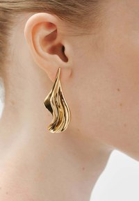 Pendiente abstracto en forma de lágrima dorada con una textura brillante, que presenta líneas onduladas y un diseño fluido, usado en la oreja.