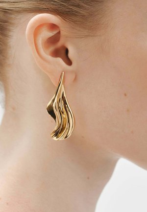 Pendiente abstracto en forma de lágrima dorada con una textura brillante, que presenta líneas onduladas y un diseño fluido, usado en la oreja.