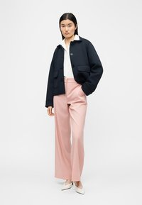 Γυναίκα που φορά μαύρο σακάκι με κουμπιά, λευκό πουκάμισο, ροζ παντελόνι με φαρδιά μπατζάκια και λευκά μυτερά ψηλοτάκουνα, στέκεται μπροστά σε ένα απλό λευκό φόντο.