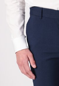 Marineblaue Stoffhosen mit glatter Textur, kombiniert mit einem weißen Hemd mit einem scharfen Bund. Hand liegt leicht auf dem Oberschenkel.