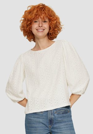 Blouse blanche texturée avec un col rond, des manches ballons et des motifs en œillets ouverts, portée avec un jean en denim bleu.