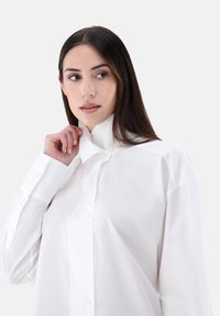 Camicia bianca con bottoni, colletto alto, maniche lunghe e vestibilità comoda. Tessuto liscio e design classico, senza motivi né accenti.