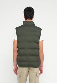 Zelená prešívaná vetrovka s vysokým golierom, ľahká tkanina. Nosená cez biele tričko, doplnená béžovými cargo nohavicami.