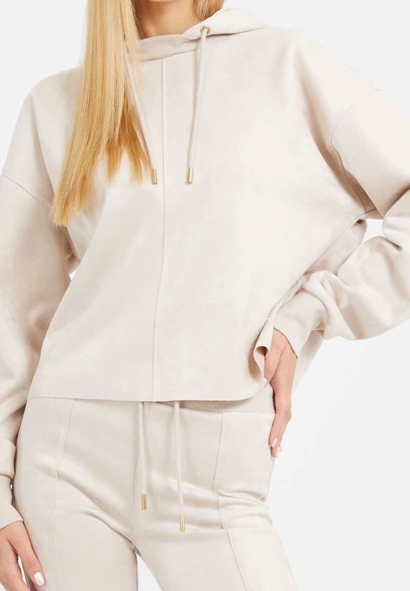 Femme portant un sweat à capuche beige clair et un pantalon de jogging assorti, main sur la hanche, cheveux blonds lisses, fond blanc.