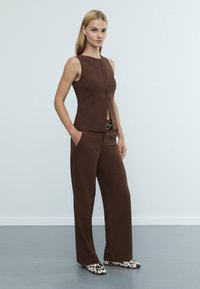 Hnědý upravený vest s kulatým výstřihem a pěti knoflíky, v kombinaci s hnědými širokými kalhotami a černobíle vzorovanými botami.