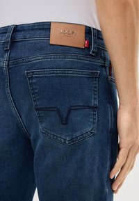 Jean en denim bleu foncé avec une étiquette en cuir portant l'inscription "JOOP! JEANS" au niveau de la taille, avec des poches arrière ornées de coutures décoratives.
