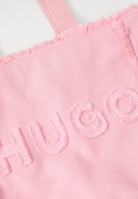 Roze stoffen draagtas met rafelige randen, met het geborduurde woord "HUGO" in bijpassend roze. Zachte textuur en eenvoudig ontwerp.