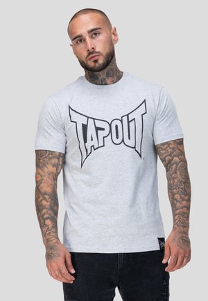 Mann mit Tätowierungen an Armen und Hals, der ein hellgraues T-Shirt mit großem schwarzem "TAPOUT"-Schriftzug trägt, vor einem schlichten Hintergrund steht.