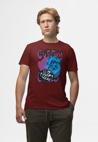 Bordová bavlněná trička s odvážným grafickým designem, na kterém je ilustrace modrého motocyklu a stylizovaný text, umístěné na fialovém pozadí.