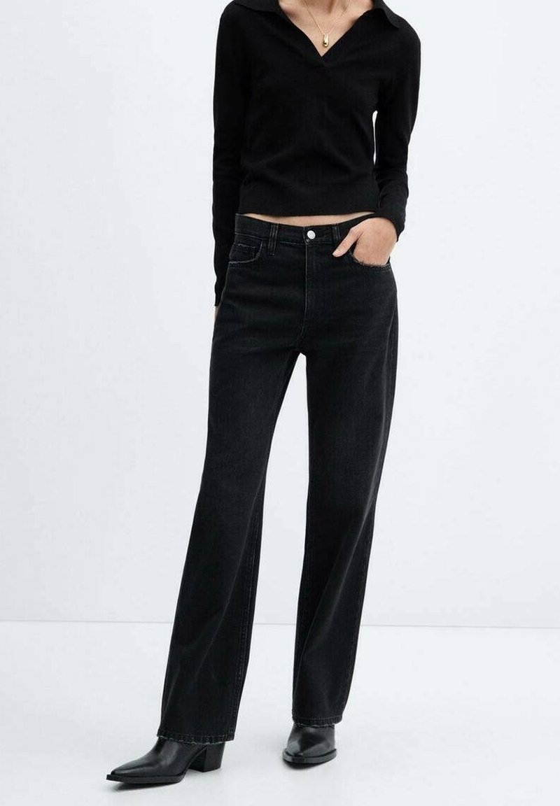 Femme portant un haut noir à manches longues et col en V, un jean noir taille haute à jambes larges, et des bottines noires à bout pointu, debout sur fond blanc.