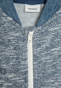 Felpa con zip blu e grigia realizzata in tessuto testurizzato, con zip bianca e un'etichetta nel colletto. Nessun motivo o dettaglio visibile.
