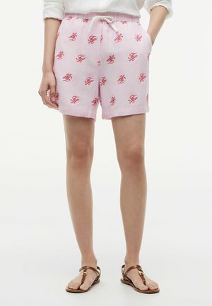 Person trägt hellrosa Shorts mit rotem Monogramm-Muster, weißem Kordelzug, weißem Oberteil und braunen flachen Sandalen, steht mit Händen in den Taschen.