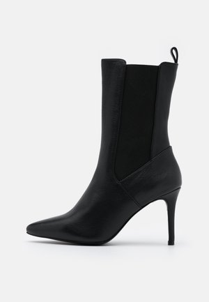 Bottine en cuir noir avec bout pointu, panneaux latéraux élastiques, languette de tirage et talon aigu fin sur fond blanc.