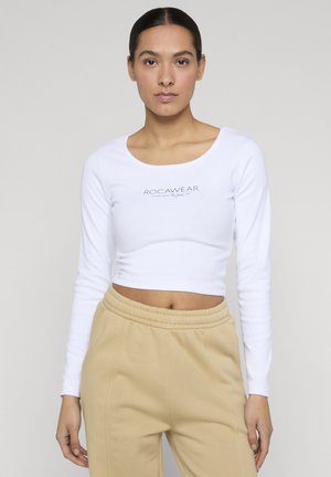Vrouw staat, draagt een wit cropped top met lange mouwen met de tekst "ROCAWEAR" en beige broek met hoge taille tegen een effen achtergrond.