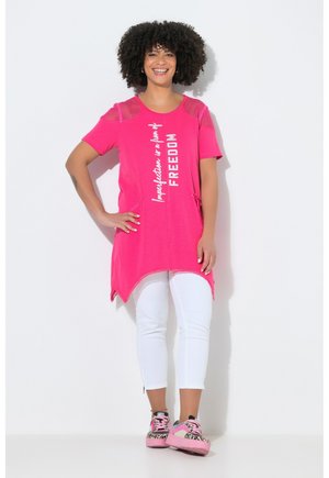 Mujer sonriente con cabello rizado viste una túnica rosa brillante con la frase "La imperfección es una forma de LIBERTAD", pantalones blancos cortos y zapatillas rosas con estampado de leopardo.