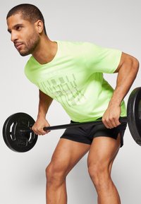 Camiseta deportiva de color verde neón con texto impreso, combinada con pantalones cortos negros. La persona está levantando una barra en una postura de entrenamiento.