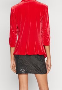 Femme portant une veste en velours rouge aux manches trois-quarts et une mini-jupe noire à sequins, vue de dos.