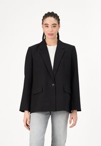 Črn blazer z enim gumbom, zarezanimi klapnicami in tremi sprednjimi žepi. Ima dolge rokave in sproščen kroj. V kombinaciji s svetlo sivimi kavbojkami.