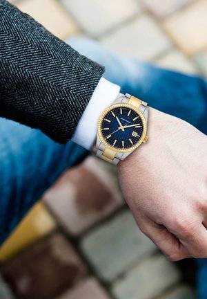 Montre en métal avec un cadran texturé bleu, des accents dorés et une fonction date. Elle possède un bracelet bicolore en argent et or, porté au poignet.