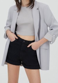 Femme portant un blazer gris clair, un crop top gris et un short noir à ourlet roulé, les mains dans les poches, devant un fond uni.