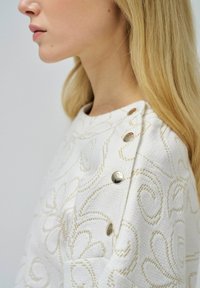 Pull blanc avec un délicat brodé floral doré et des boutons-pression argentés à l'épaule, mettant en avant une texture de tissu travaillée.