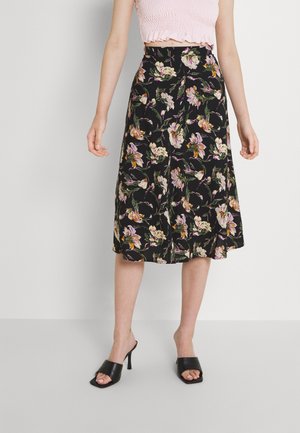 Femme portant une jupe midi noire à imprimé floral coloré, un top court rose clair et des sandales à talons ouvertes noires, debout contre un mur blanc.