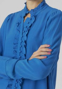 Niebieska bluzka z zapięciem na guziki z falbankami, długimi rękawami i teksturowanym materiałem. Guziki są wyraźnie widoczne wzdłuż placki.