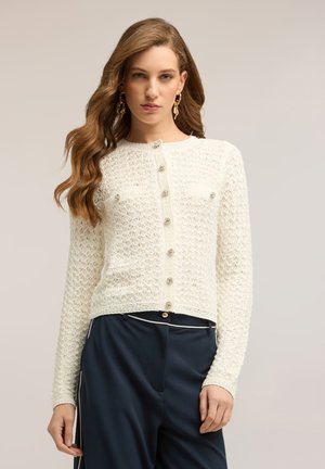 Kobieta z długimi falującymi włosami, nosząca złote kolczyki, biały teksturowany rozpinany sweter oraz granatowe spodnie z białym przeszyciem.