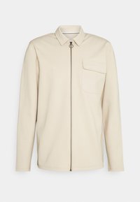 Giacca beige con zip e colletto, maniche lunghe e taschino sul petto. Realizzata in tessuto strutturato con zip a anello argentato.