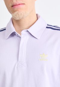 Svijetloplavi polo majica s ovratnikom, sadrži tri tamnoplava pruga na ramenima i mali žuti Adidas logo na prsima.
