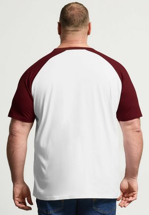 Hombre visto desde atrás con una camisa blanca con mangas raglán burdeos y vaqueros azules sobre un fondo liso.