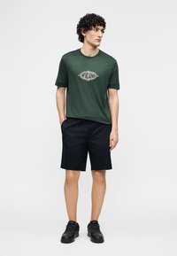 Άνδρας που φοράει σκούρο πράσινο μπλουζάκι με λογότυπο, μαύρο σορτς και μαύρα αθλητικά παπούτσια, στέκεται με τα χέρια στις τσέπες, κοιτάζοντας προς τα πλάγια.