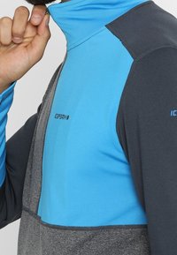 Pulóver de media cremallera en azul y gris, con una textura suave, colores contrastantes y un cuello alto para mayor abrigo. Logo visible.