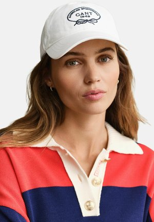 Femme aux cheveux châtain clair portant une casquette blanche GANT et un pull à col rouge, bleu marine et blanc avec des boutons dorés.